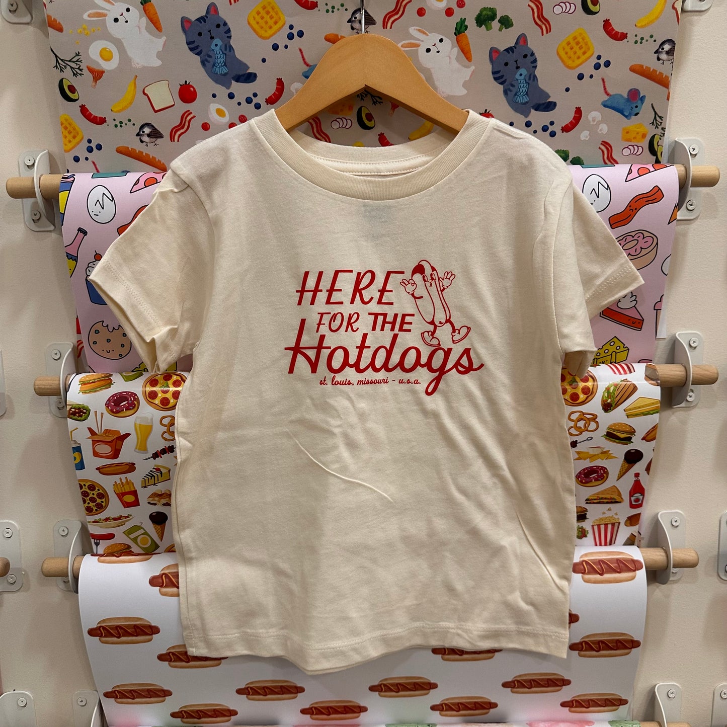 Beige t-shirt with 'Here for the Hotdogs' text on a hanger against a colorful food-themed wallpaper.