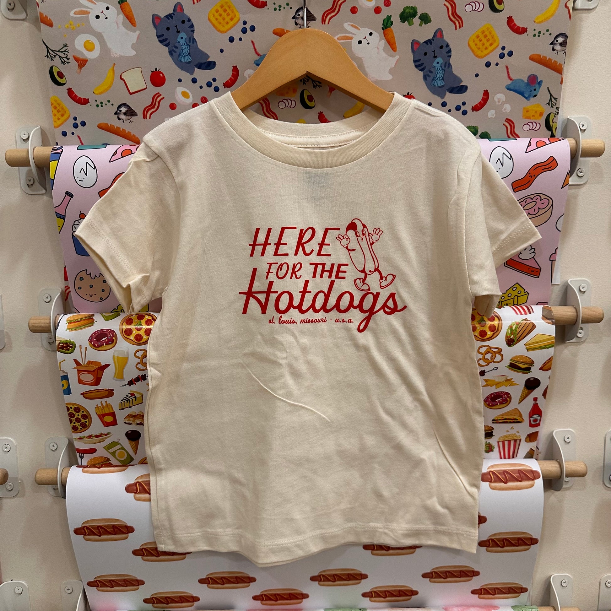 Beige t-shirt with 'Here for the Hotdogs' text on a hanger against a colorful food-themed wallpaper.