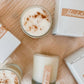 White candles in frosted glass with a sprinkle of cinnamon on top, labeled with "Horchata" on a wooden table, pictured next to white boxes it comes packaged in 