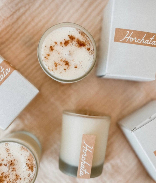White candles in frosted glass with a sprinkle of cinnamon on top, labeled with "Horchata" on a wooden table, pictured next to white boxes it comes packaged in 