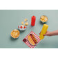 Hot dog shaped plate with mustard, ketchup, and chips on a checkered napkin with a hand holding a yellow mustard bottle.