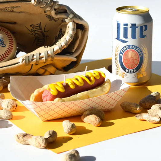 Hot dog candle with mustard in paper boat for display, along with a baseball glove, beer can, and peanuts