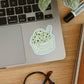 A die-cut sticker of a mint chip ice cream cup with the text 'Ice cream is ALWAYS a good idea' applied to the palm rest of a silver laptop on a wooden desk, surrounded by a plant, glasses, and a marker.