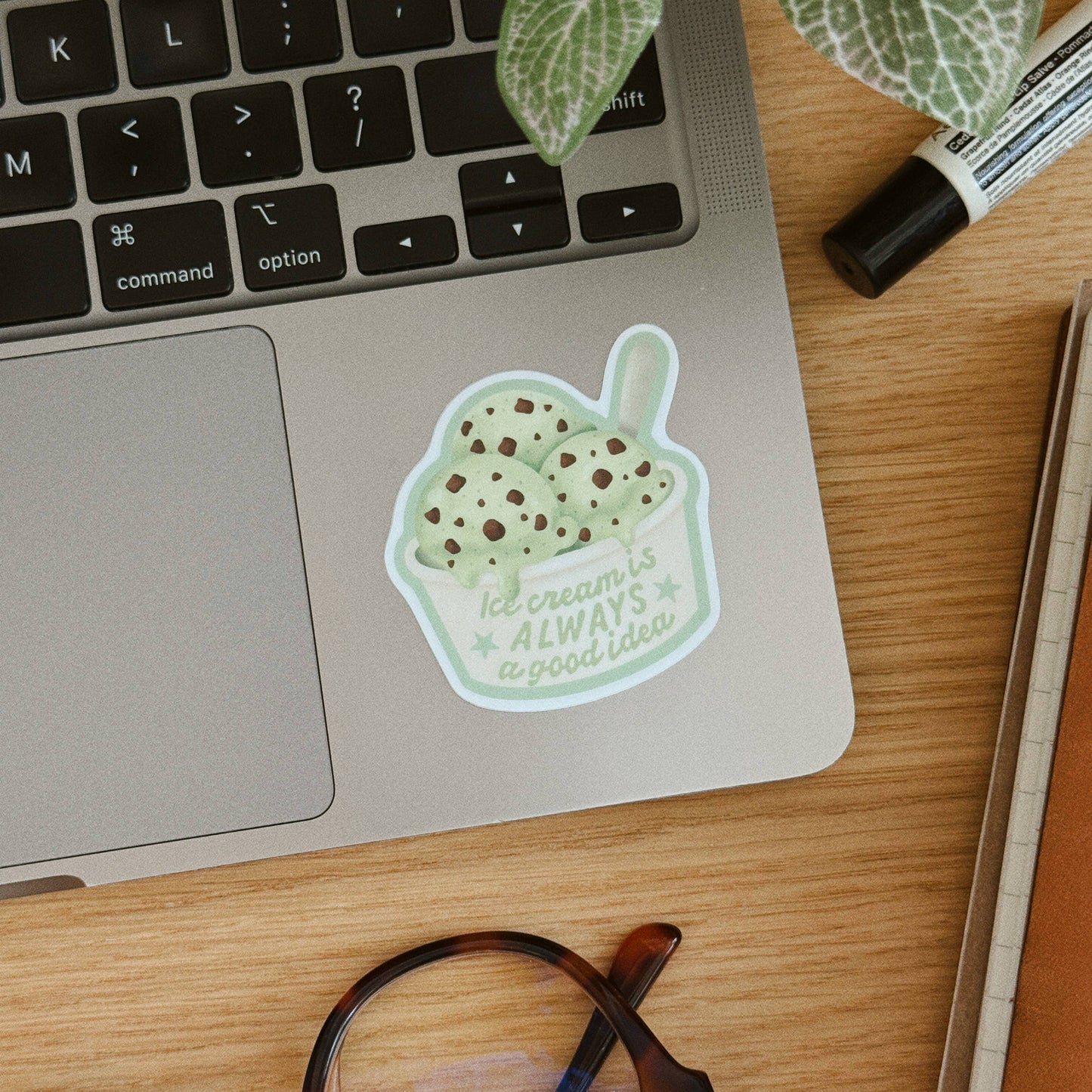 A die-cut sticker of a mint chip ice cream cup with the text 'Ice cream is ALWAYS a good idea' applied to the palm rest of a silver laptop on a wooden desk, surrounded by a plant, glasses, and a marker.