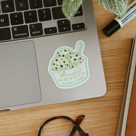 A die-cut sticker of a mint chip ice cream cup with the text 'Ice cream is ALWAYS a good idea' applied to the palm rest of a silver laptop on a wooden desk, surrounded by a plant, glasses, and a marker.