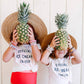 Two children holding pineapples in front of their faces, wearing matching shirts and hats.