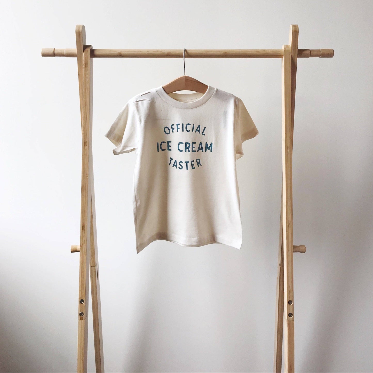 Beige t-shirt with 'Official Ice Cream Taster' text on a wooden rack against a white background