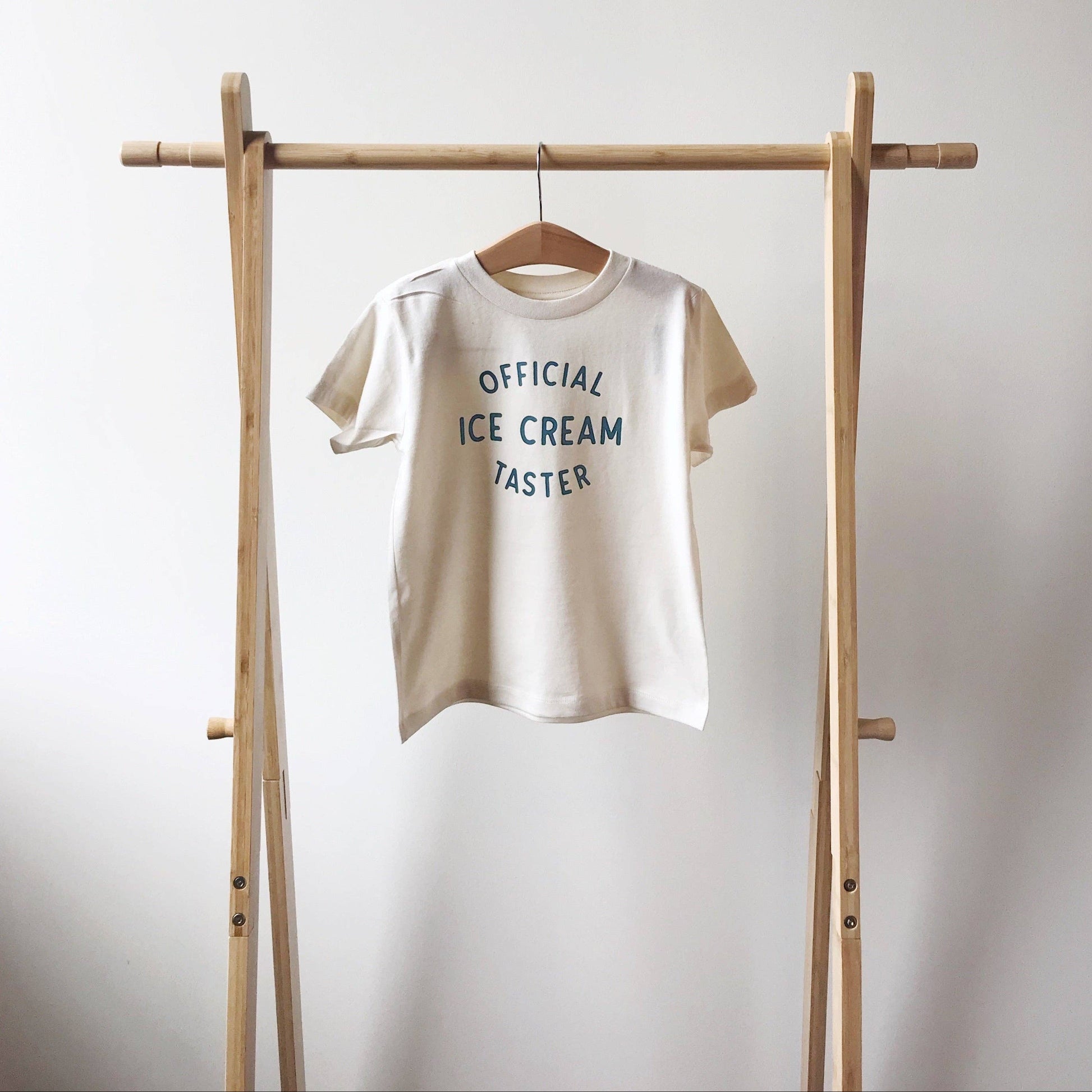 Beige t-shirt with 'Official Ice Cream Taster' text on a wooden rack against a white background
