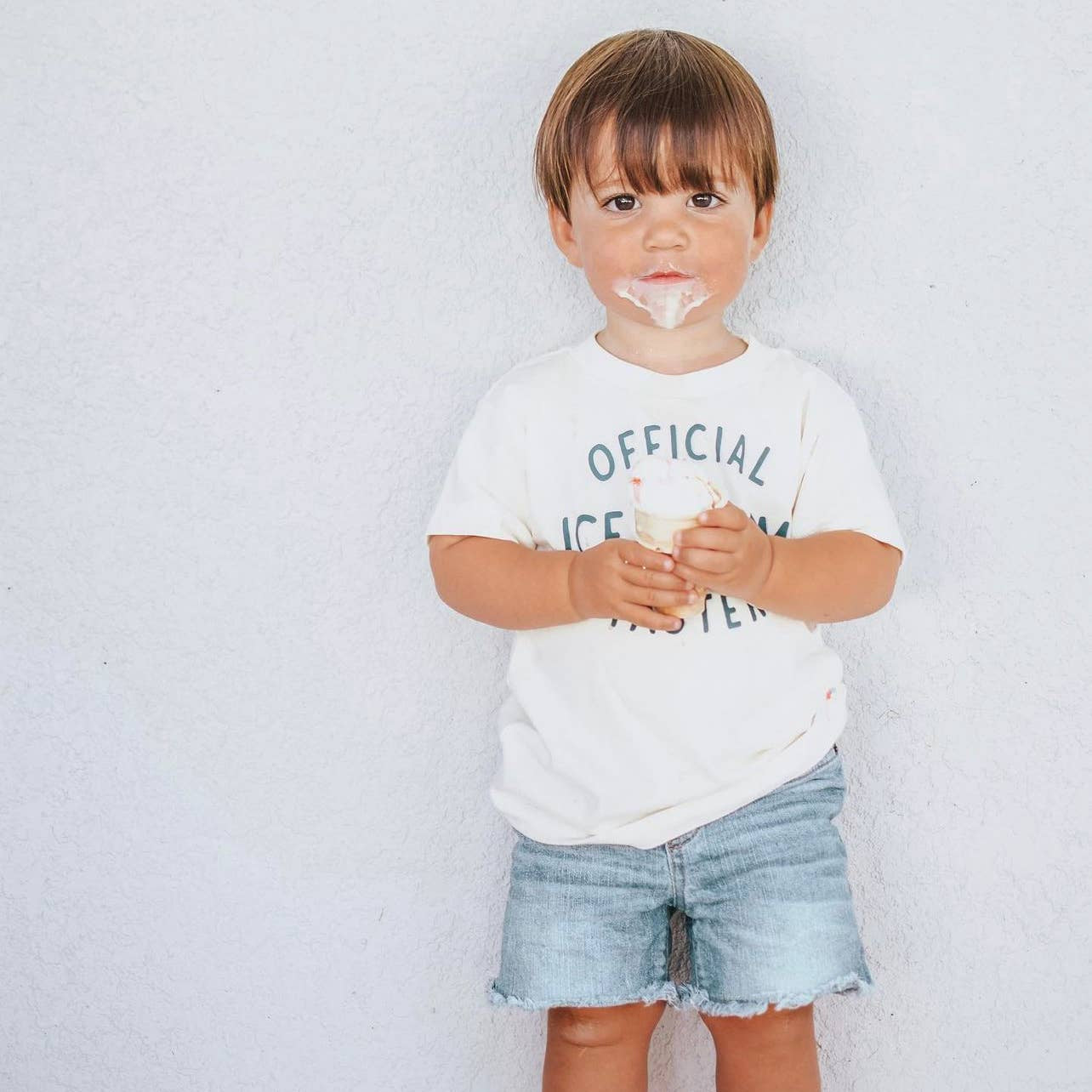 Child wearing a white t-shirt with text and denim shorts, holding an ice cream cone against a plain background