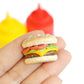 Photo of single tiny cheeseburger magnet resting on a person's fingertips with yellow mustard bottle and red ketchup bottle in the background.