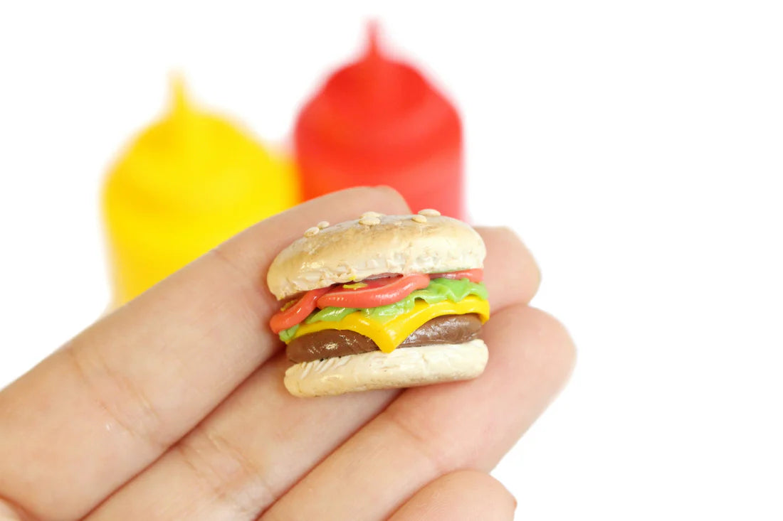 Photo of single tiny cheeseburger magnet resting on a person's fingertips with yellow mustard bottle and red ketchup bottle in the background.