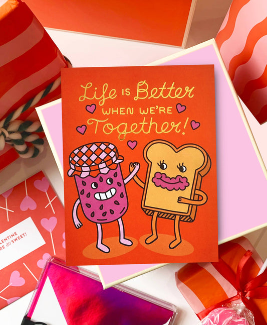 red greeting card with gold foil text that reads "Life is better when we're together" Has an illustration of a jam jar holding hands with a slice of toast