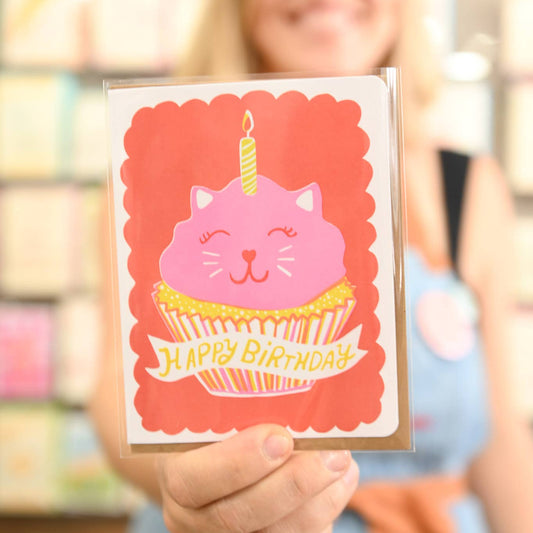 Person holding a birthday card with a pink cat on a cupcake, blurred background