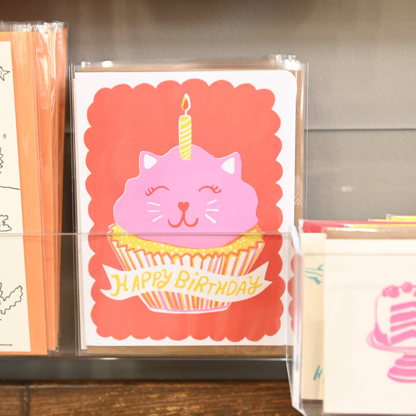 Collection of greeting cards with a pink cat in a cupcake design on a shelf.