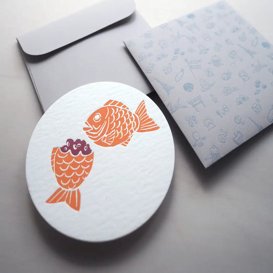 round white notecards with red bean filled taiyaki design on it. Envelope is light grey with various illustrations of notable landmarks, attractions, food and drink from Japan 
