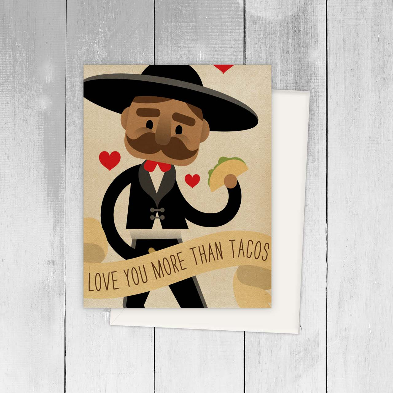 Card with a cartoon mariachi holding a taco and a banner saying 'Love you more than tacos' on a wooden surface.