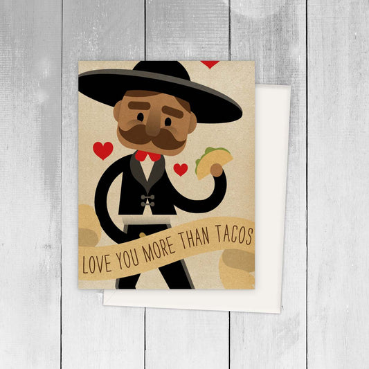 Card with a cartoon mariachi holding a taco and a banner saying 'Love you more than tacos' on a wooden surface.