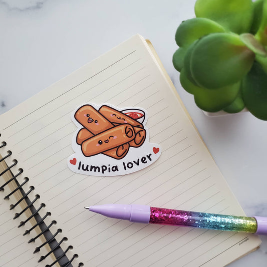 A sticker with 5 lumpias and sweet & sour sauce on it, with text at bottom that reads "lumpia lover" Placed here with a lined notebook and rainbow pen.