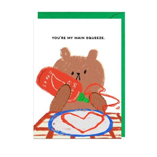 greeting card with a bear at a table, squeezing a bottle of sriracha and making a heart on a plate. Text reads "You're my main squeeze" 