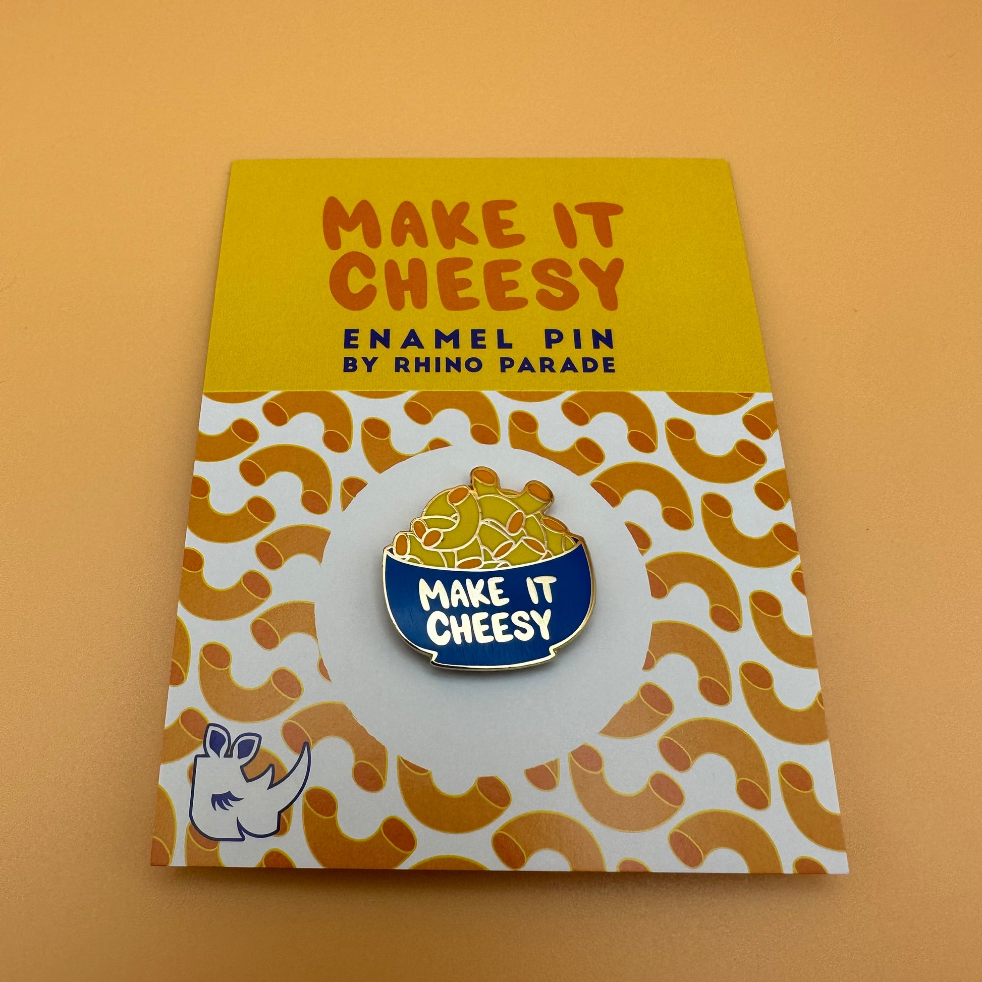 Bowl of mac and cheese enamel pin that reads "make it cheesy." On a card backing that has a patterned image of macaroni moodles.