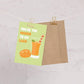 Greeting card with a cut mango and glass of mango lassi on a green background, hung on string with clips. Text reads "You're the mango to my lassi" 