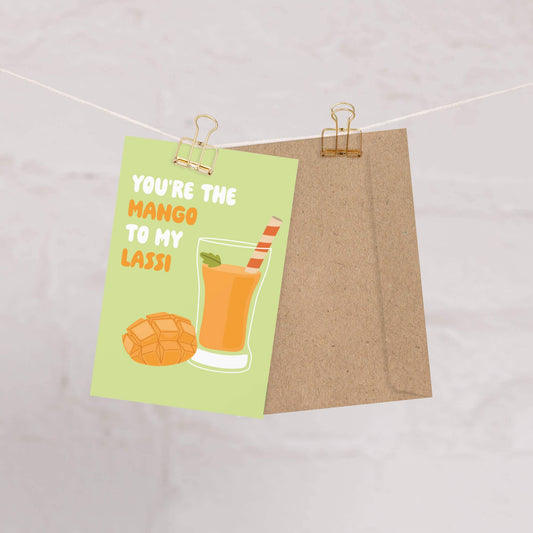 Greeting card with a cut mango and glass of mango lassi on a green background, hung on string with clips. Text reads "You're the mango to my lassi" 