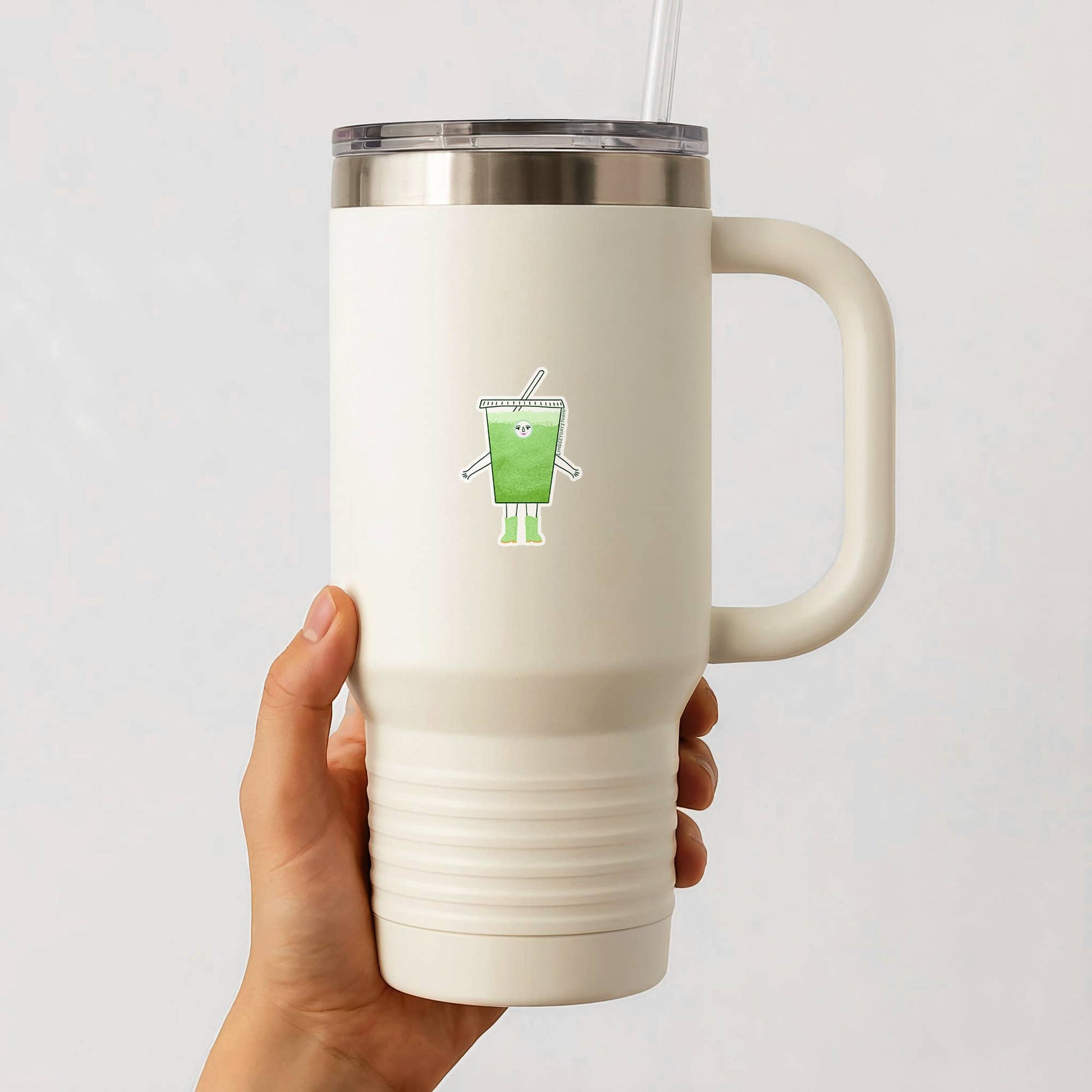 White insulated mug with a iced matcha cartoon character sticker holding a drink, held by a hand against a light gray background