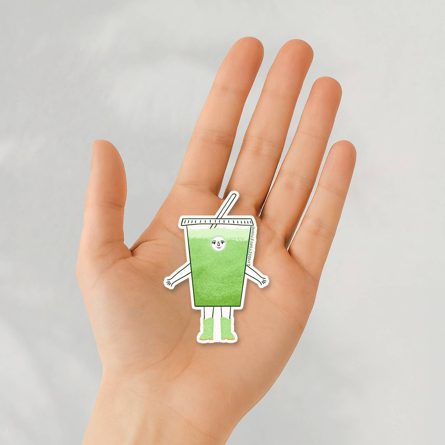 hand holding a sticker of an iced matcha character 
