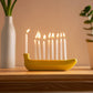 Yellow banana-shaped candle holder/menorah with lit candles on a wooden surface, with a vase and plant in the background.