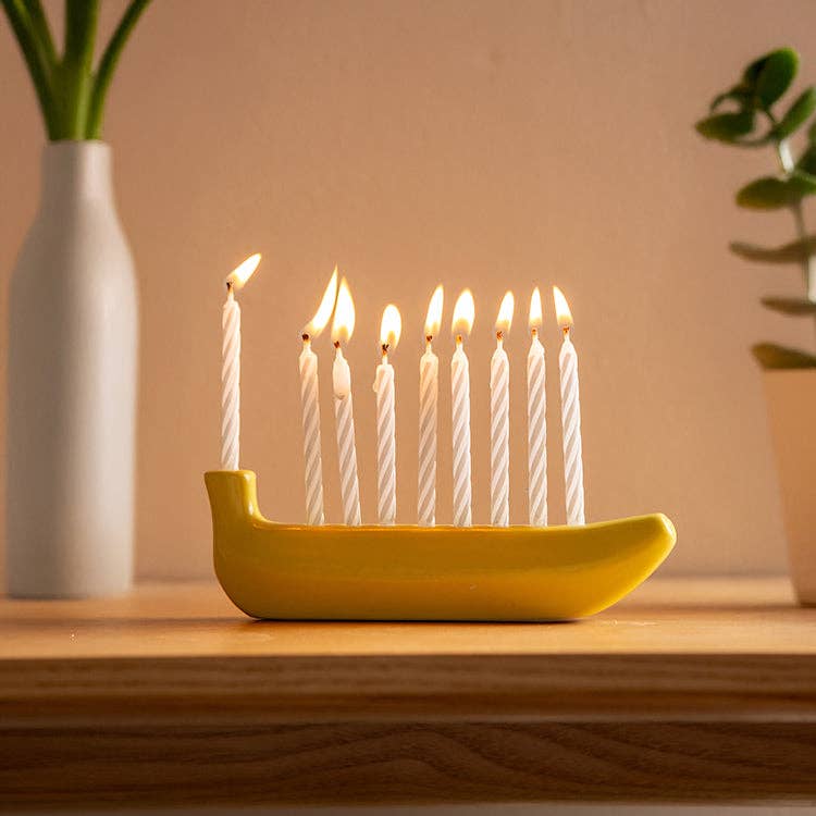 Yellow banana-shaped candle holder/menorah with lit candles on a wooden surface, with a vase and plant in the background.