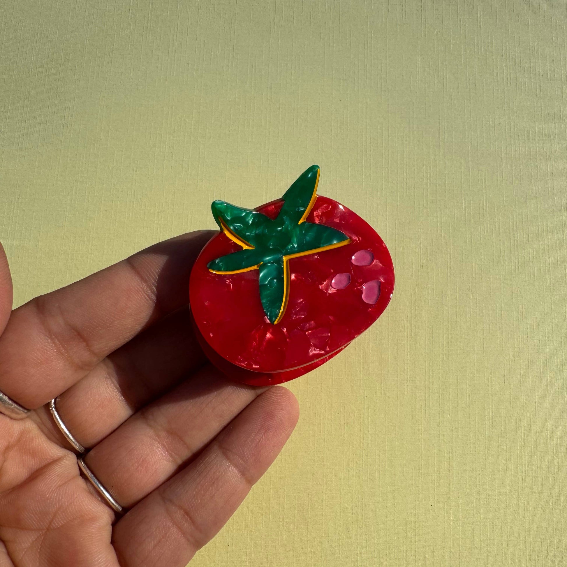 Red tomato-shaped mini hair claw with green leaves held in a hand against a yellow background