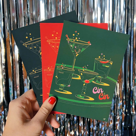 Hand holding 3 different colored Cin Cin prints in black, red and green. Cin Cin print has different cocktails with detailed lined drawing.