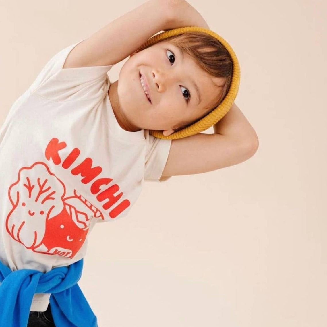 Happy toddler wearing yellow hat and Kimchi Tee Shirt. Kimchi Tee -- says "Kimchi" across the chest with a cabbage next to a pickling jar, all in red ink.