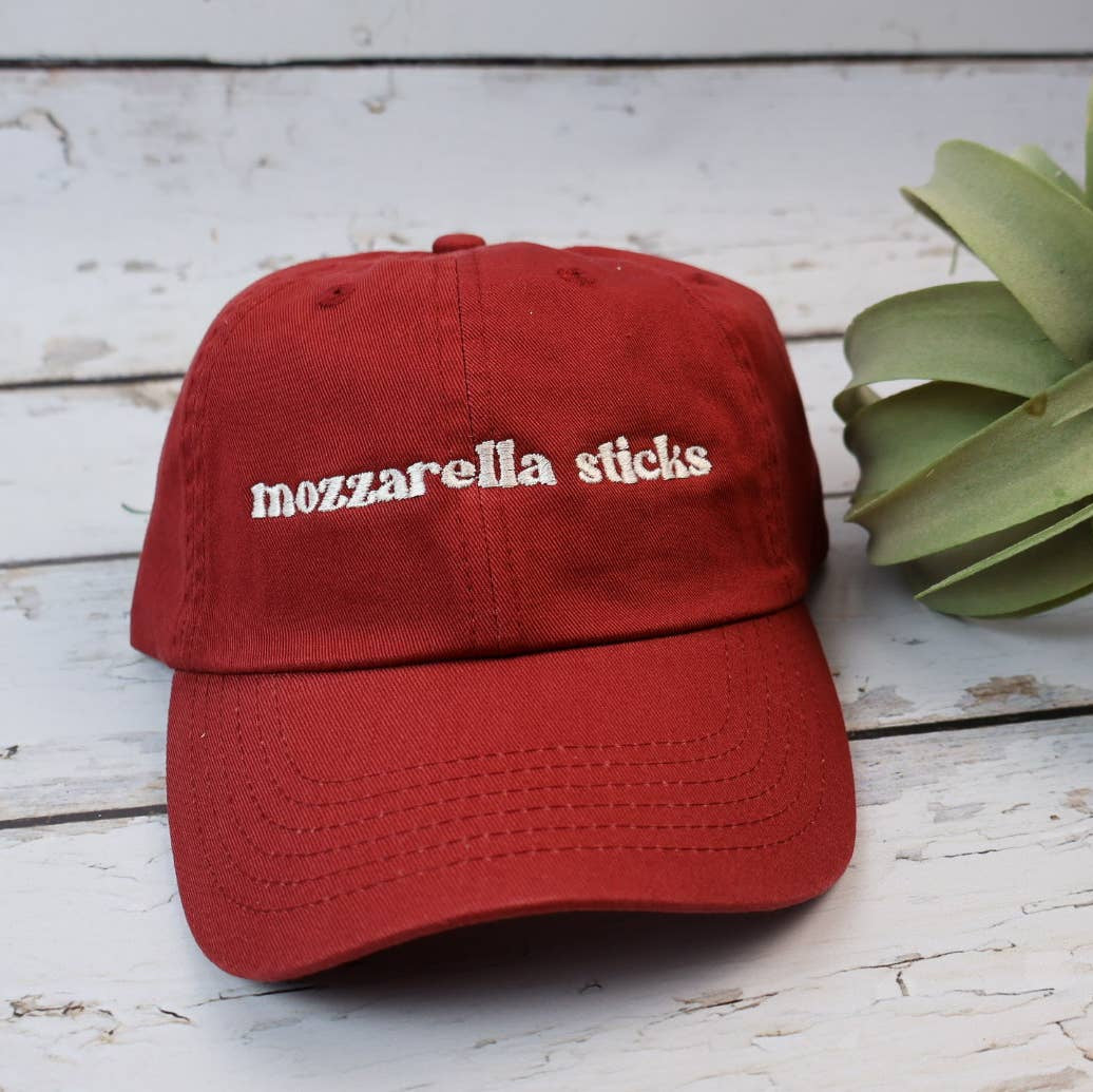 A red baseball cap with 'mozzarella sticks' embroidered in white on the front, styled on a white distressed wood background next to a green succulent plant.
