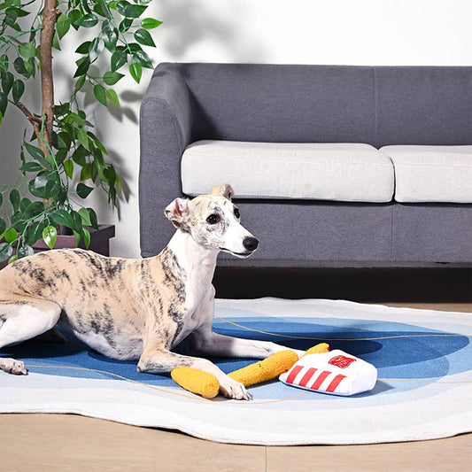 Dog lying on a blue mat with a plush mozzarella sticks toy in a living room setting