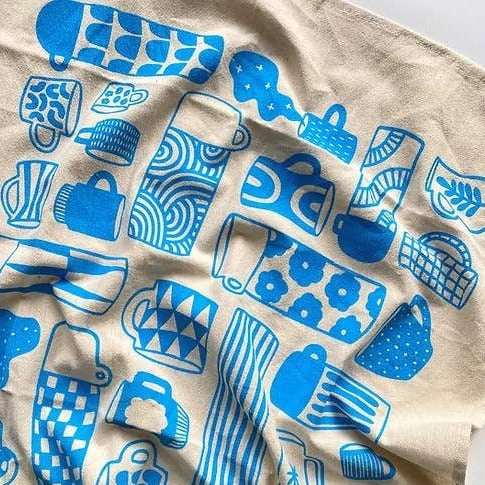 Kitchen towel with a variety of coffee cups, mugs and thermoses that are decorated with different retro/geometric decorative designs. Printed in blue ink on natural colored fabric.