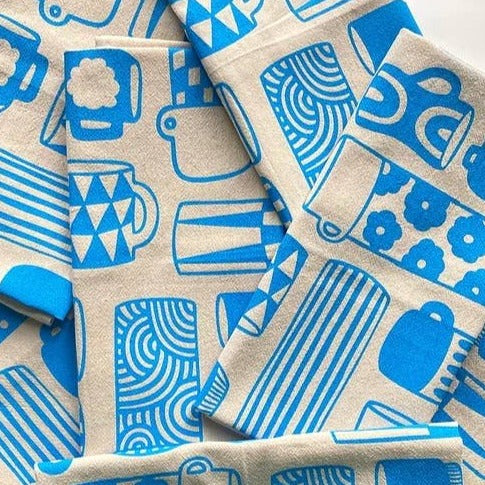 Kitchen towel with a variety of coffee cups, mugs and thermoses that are decorated with different retro/geometric decorative designs. Printed in blue ink on natural colored fabric.