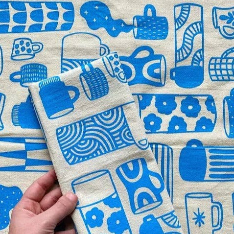 Kitchen towel with a variety of coffee cups, mugs and thermoses that are decorated with different retro/geometric decorative designs. Printed in blue ink on natural colored fabric.