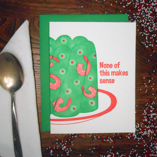 Card with green and pink jello design with shrimp and olives inside and text 'None of this makes sense' on a wooden surface with a spoon and confetti.