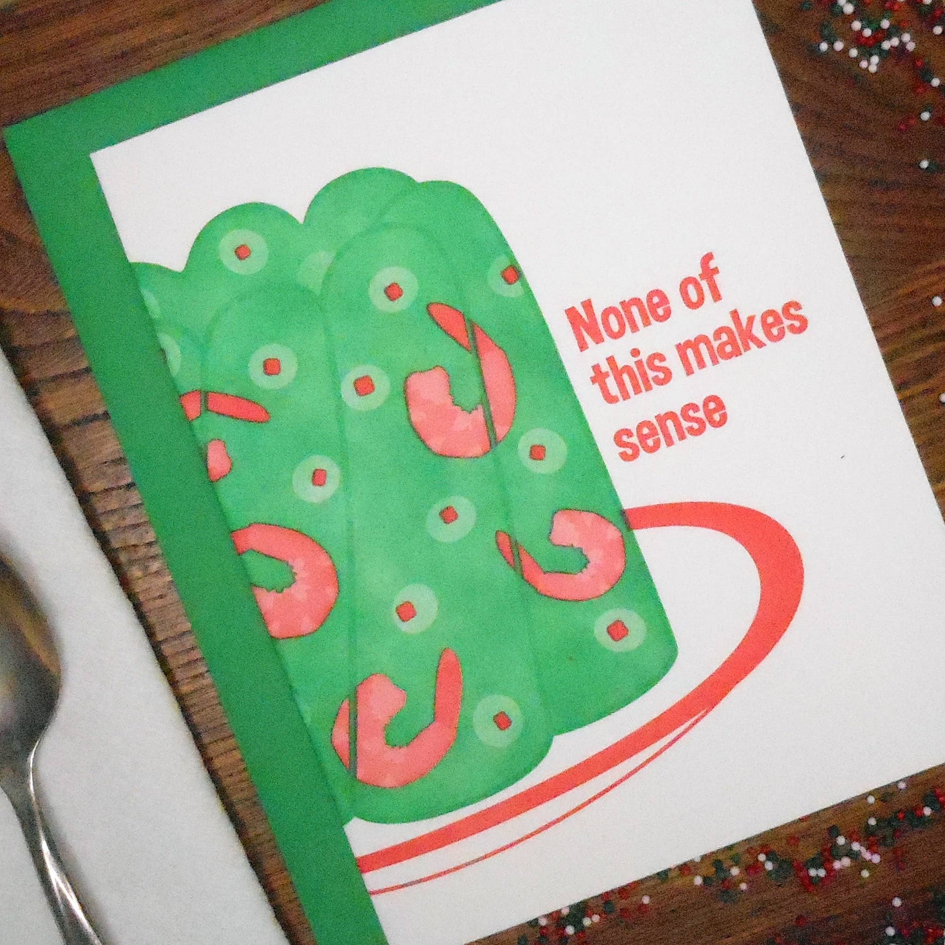 Card with green jello design with shrimp and olives inside, text 'None of this makes sense', on a wooden surface.