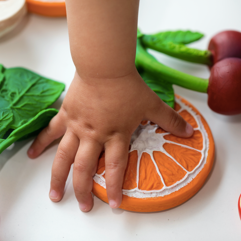 baby hand reaching for orange slice baby teething toy. Other teethers pictured -- radish, cherry, mushroom and apple 