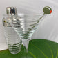 Clear martini glass with a cocktail shaker and an olive cocktail pick on a green surface