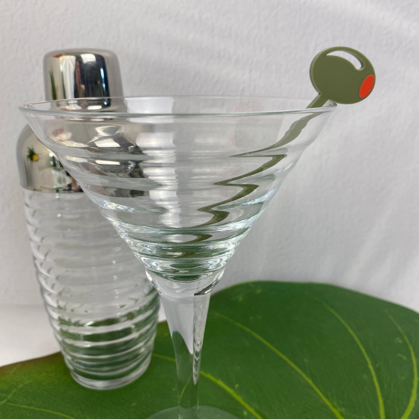 Clear martini glass with a cocktail shaker and an olive cocktail pick on a green surface