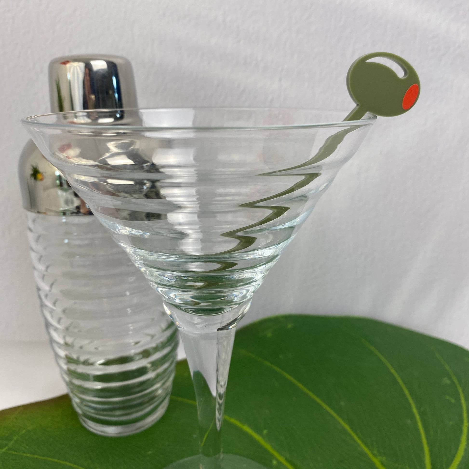 Clear martini glass with a cocktail shaker and an olive cocktail pick on a green surface