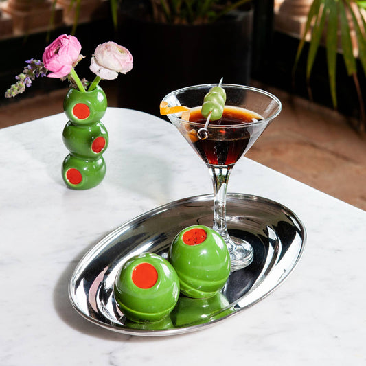 Stuffed green olive salt and pepper shakers on a silver tray next to a cocktail in a martini glass.
