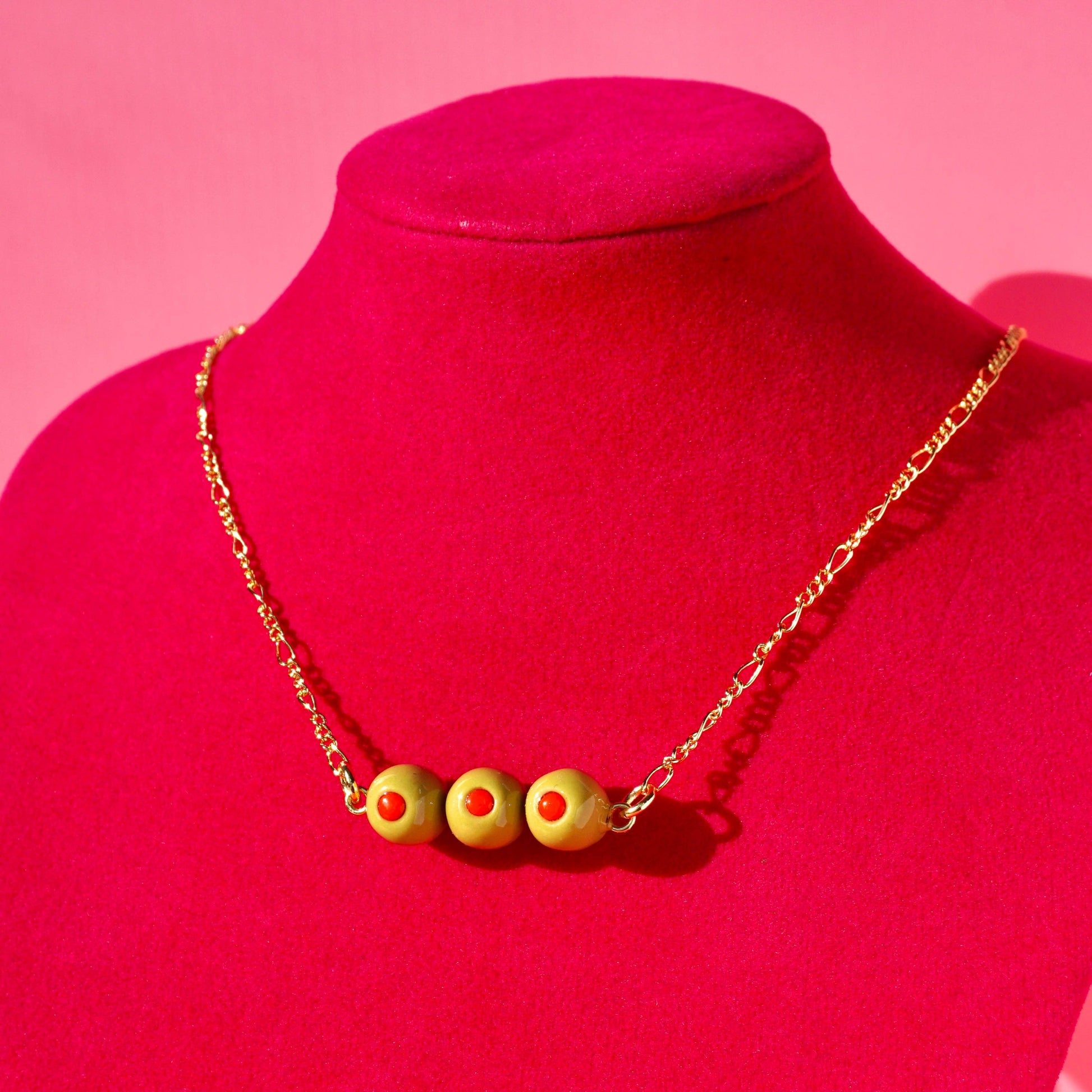 Necklace with gold chain and set of 3 stuffed green olive charms on a red mannequin against a pink background