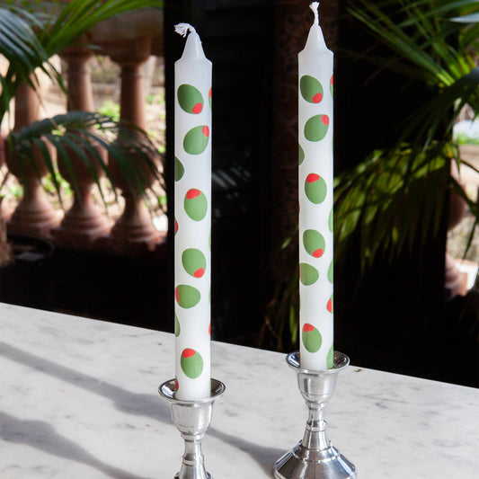Two candles with stuffed green olive design in silver candle holders on a marble surface with a blurred natural background.