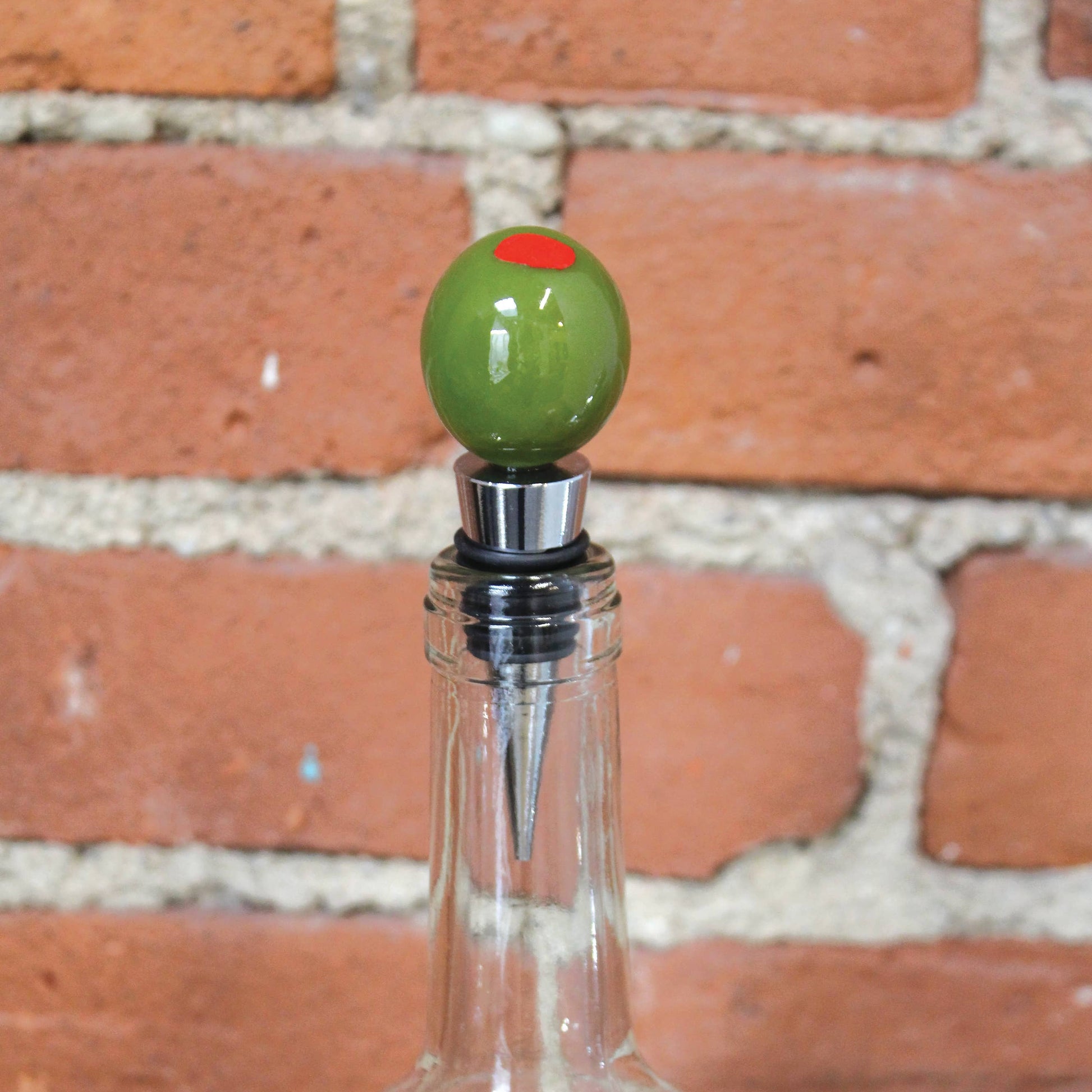 Clear bottle with a green olive-shaped stopper against a brick wall.