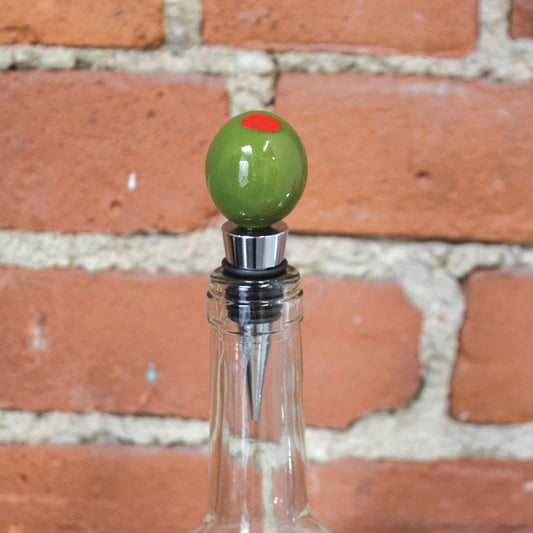 Clear bottle with a green olive-shaped stopper against a brick wall.
