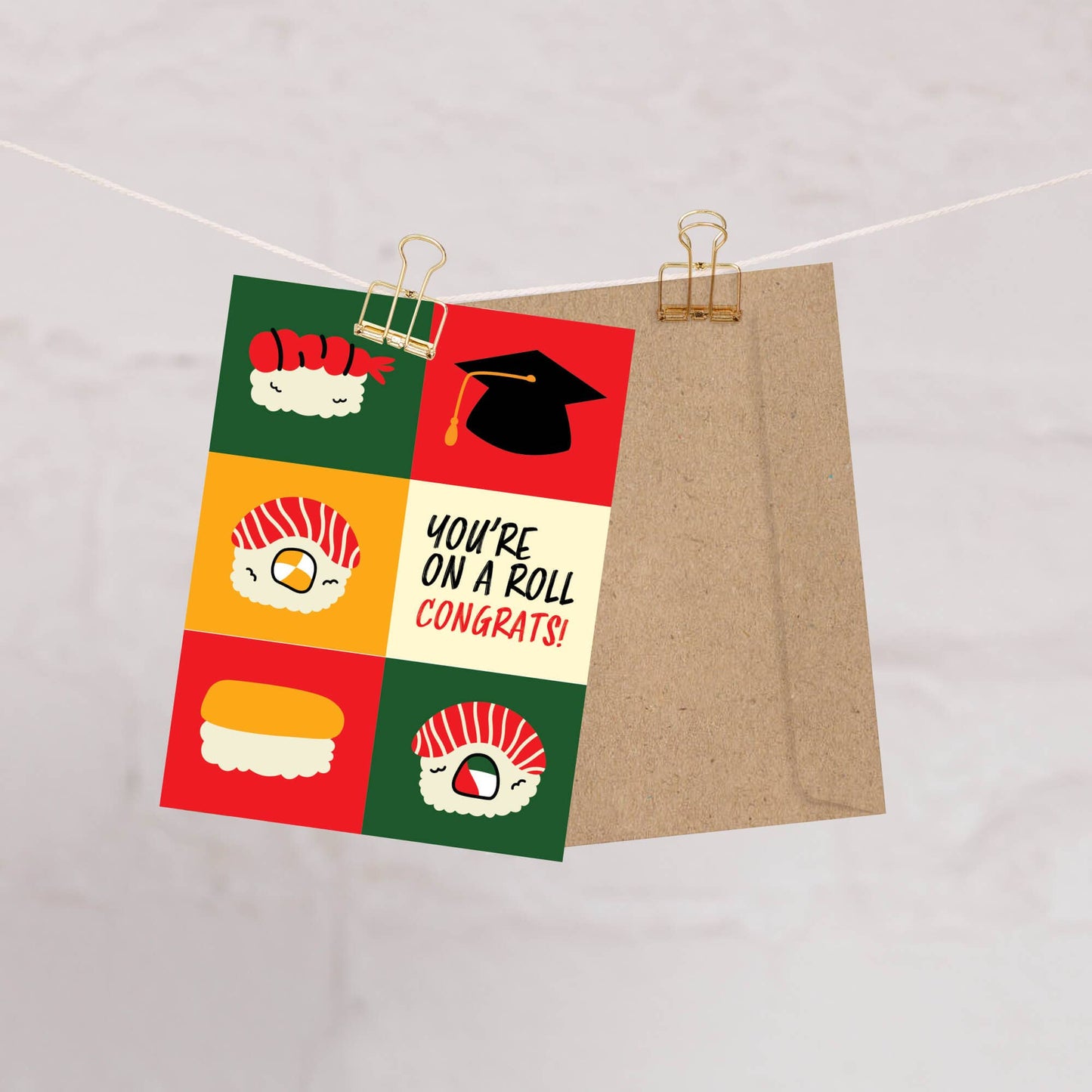 Greeting card with colorful sushi-themed design and 'You're on a roll, congrats!' text, held open by a paper clip against a light background.